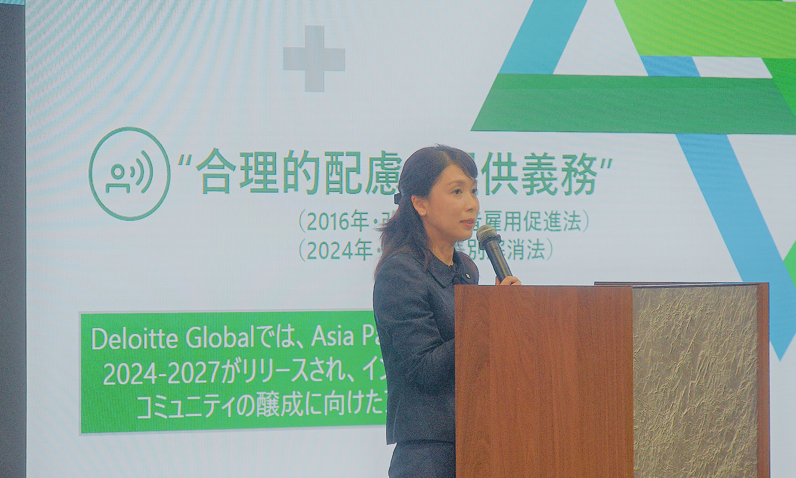 ALTはじまり。司会者台の前に、マイクを持って講演をするスーツ姿の青野様。背景にはプレゼン資料が映し出されたスクリーンが写っている。ALTおわり