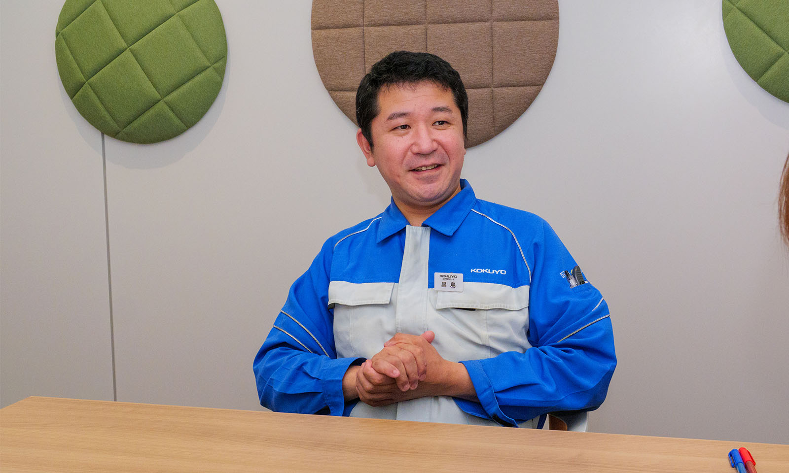 ALTはじまり。壁に黄緑やブラウンの丸いファブリックオブジェが貼り付けられた会議室で、黒髪短髪の男性が両の手のひらをみぞおちのあたりで合わせながら笑顔で語っている。左胸にKOKUYOのロゴが入った青と白の制服を着用している。ALTおわり。