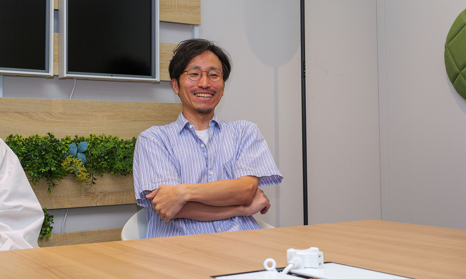 ALTはじまり。ナチュラルな木の温もりと緑の差し色が映える会議室のモニター前で、細いフレームのメガネをかけた男性が腕を軽く組みながら笑顔で語っている。青と白のストライプ柄がさわやかな印象を与える襟付きのシャツを着ている。ALTおわり。