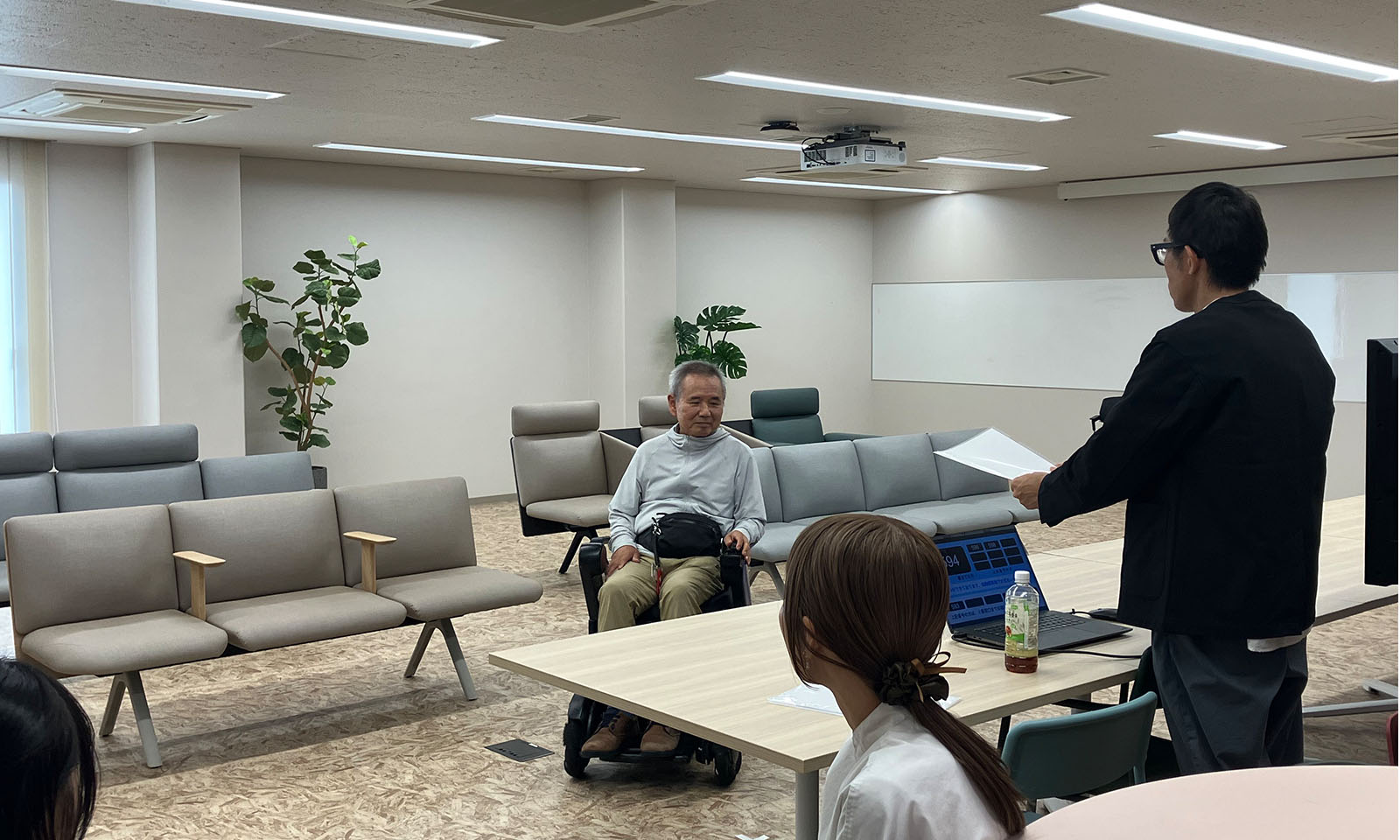 ALTはじまり。開発したロビーチェアーが4つ並べられた白を基調とする空間。車椅子に乗った白髪混じりの男性を前に、黒い長そでを着たメガネの男性が書類を片手に説明をしている。手前には髪の毛を後ろで一つに束ねた白服の女性の姿があり、2人の様子を見守っている。ALTおわり。