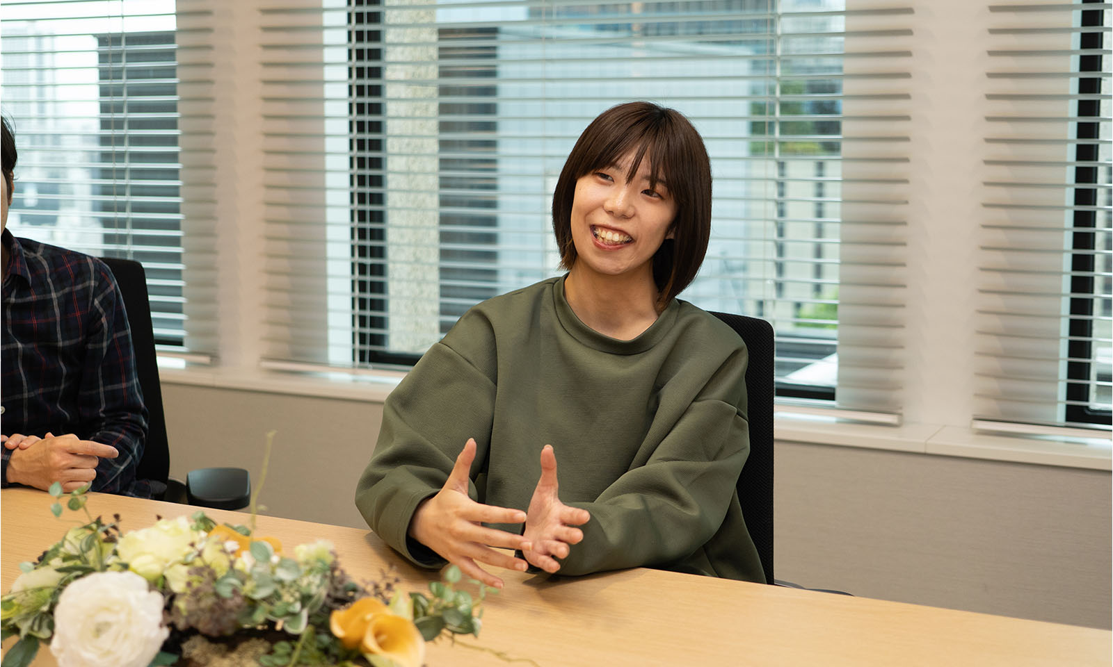 ALTはじまり。ゆったりしたモスグリーンのニットシャツを着ているコクヨの高木さん。中央に花やグリーンが飾られた会議室のテーブルの一角で、手を広げながら製品について笑顔で説明している。ALTおわり