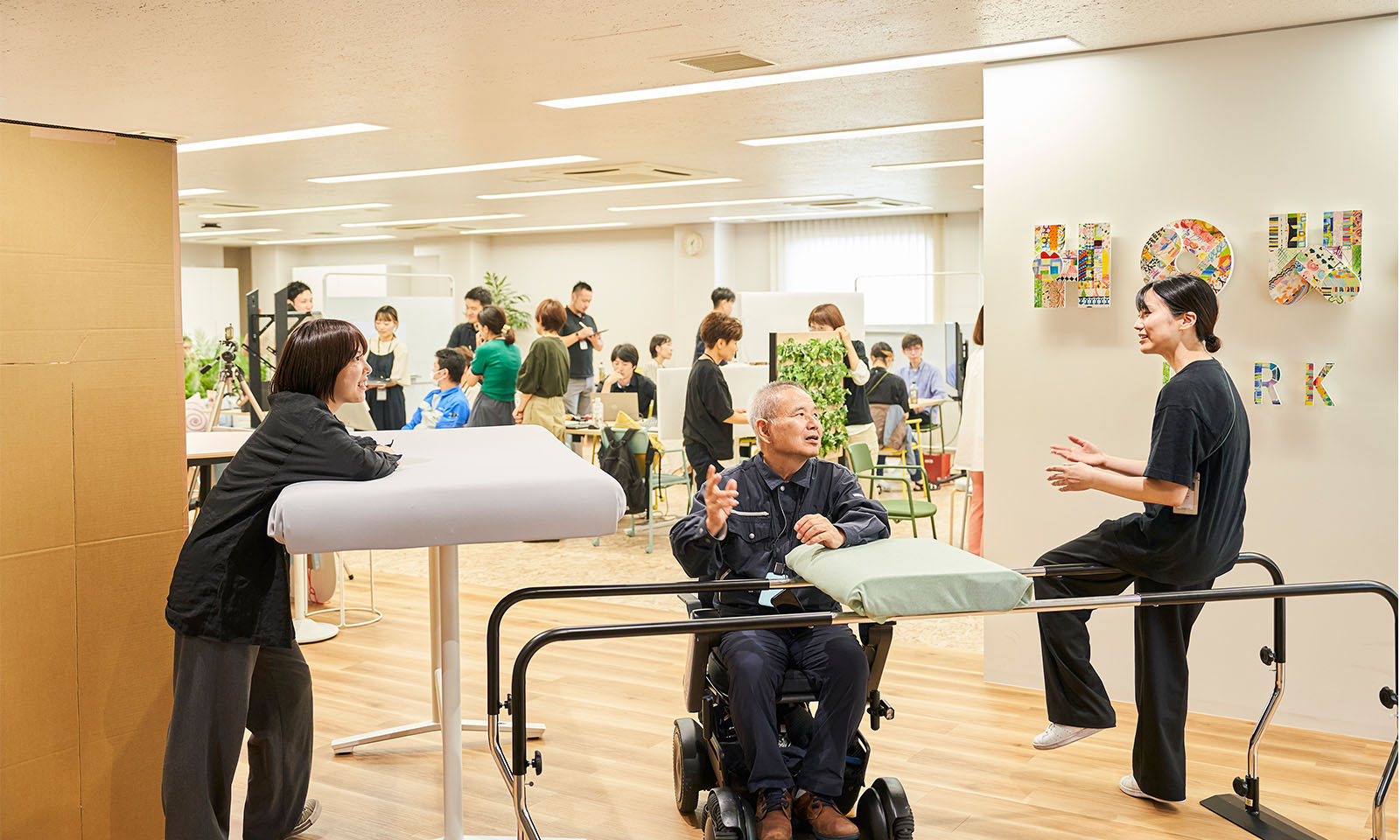 ALTはじまり。ワークショップの様子。広い部屋に20人ぐらいの人がまばらに集まって話している。部屋の中央には車椅子に座った黒いジャージ上下を着た宮原さんがいて、黒シャツと黒パンツを着て手すりに腰を下ろす高木さんと話している。少し離れたところには入テーブルがあり、黒のシャツとゆったりしたパンツ姿の小林さんが、立って肘をついている。ALTおわり。