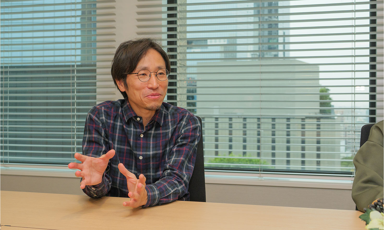 ALTはじまり。会議室のテーブルに着き、身振りを交えて笑顔で話す靏崎さん。グリーンとブルーのチェックシャツを着て、眼鏡をかけている。ALTおわり。