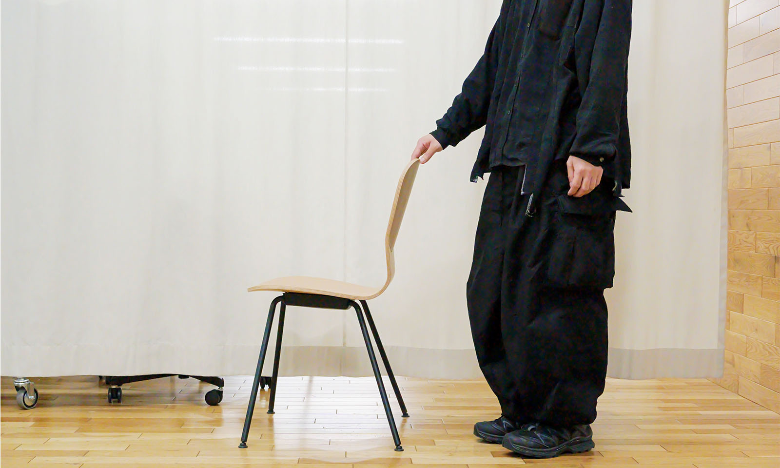 ALTはじまり。イスの背もたれに手をかけている様子を真横から撮影。S字の曲線を描くスタイリッシュな背もたれと、すらりと伸びる黒い脚の形状がよく分かる。イスに触れているのは全身を黒でコーディネートした男性で、肩から下の半身のみが映っている。ALTおわり。