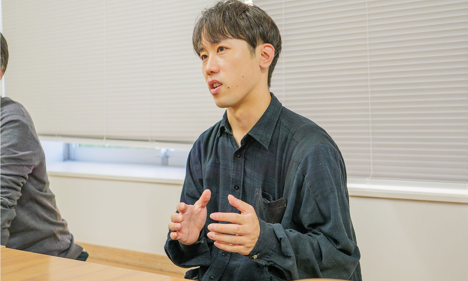 ALTはじまり。ブラインドを下げた窓がある空間。前髪を下ろした短髪の男性が両手を少し広げて、真剣な面持ちで喋っている。着用しているのは、一見すると黒一色に見える襟付きの長そでだが、よく見るとチェック模様のデザインが施されている。ALTおわり。