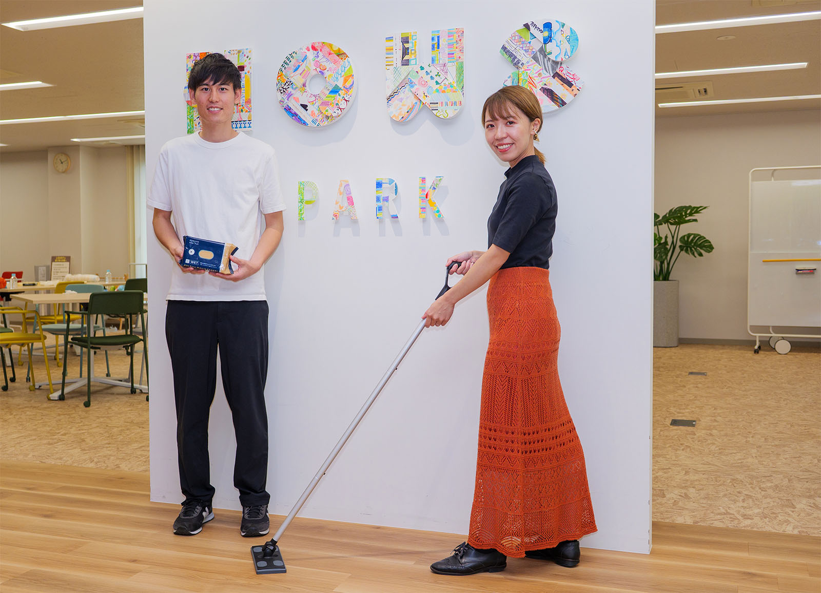 <ALT始まり>インタビュイー1人目の大枝さんが「職場の靴跡汚れさえも落とせるフローリングワイパー用ウェットシート」を持って左側に、インタビュイー2人目の本間さんが「文具のコクヨが本気で消しやすさにこだわった軽い力でスルッと落とせるフローリングワイパー」を床にかけるポーズを取って右側に立っている。HOWS PARKの入口の前にいる。<ALT終わり>