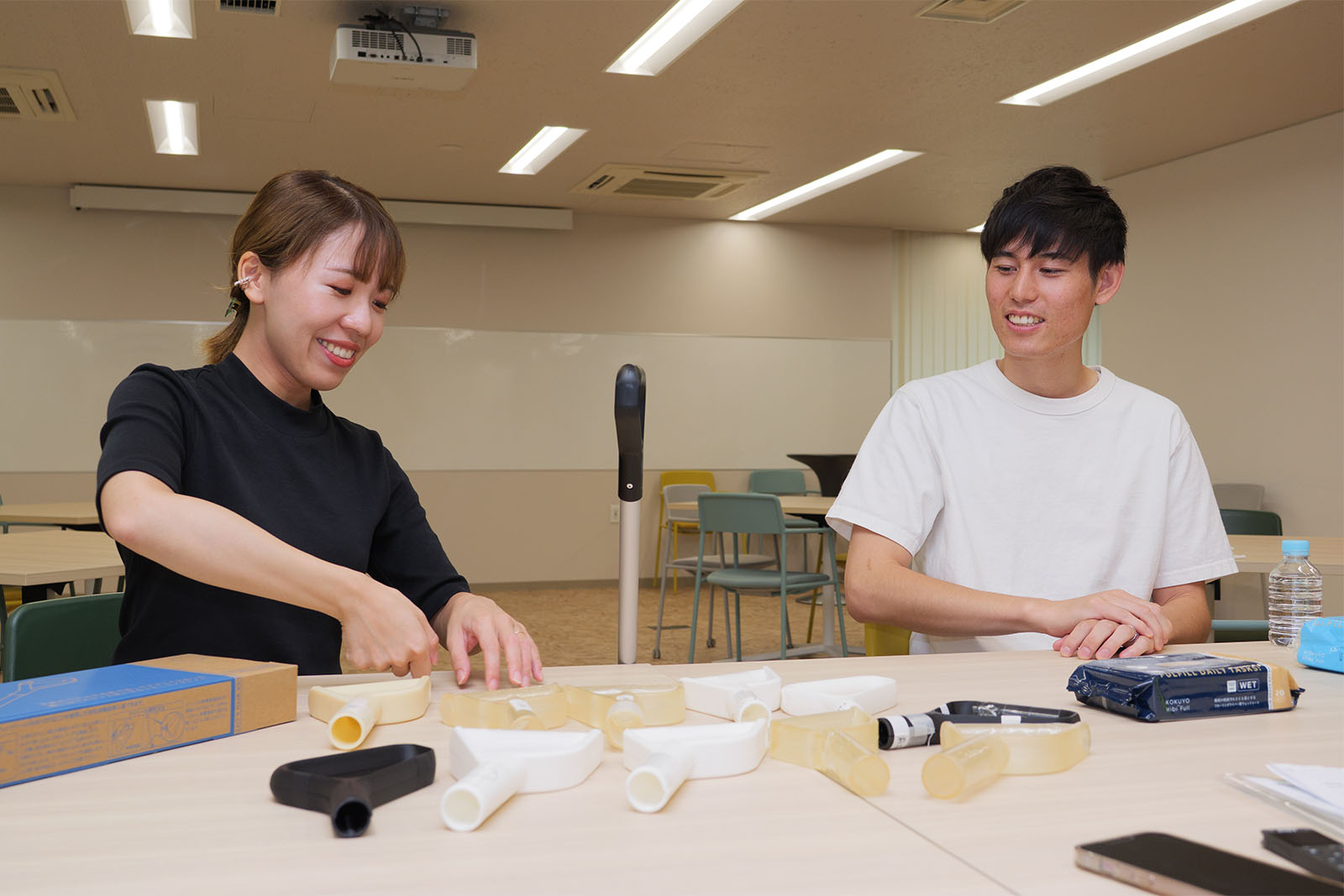 <ALT始まり>インタビュイーの本間さんが、持ち手の形状の説明をしている。机上にはワイパーの持ち手の試作品が10点置いてある。本間さんは黒いシャツを着ている。隣に大枝さんがいる。<ALT終わり>