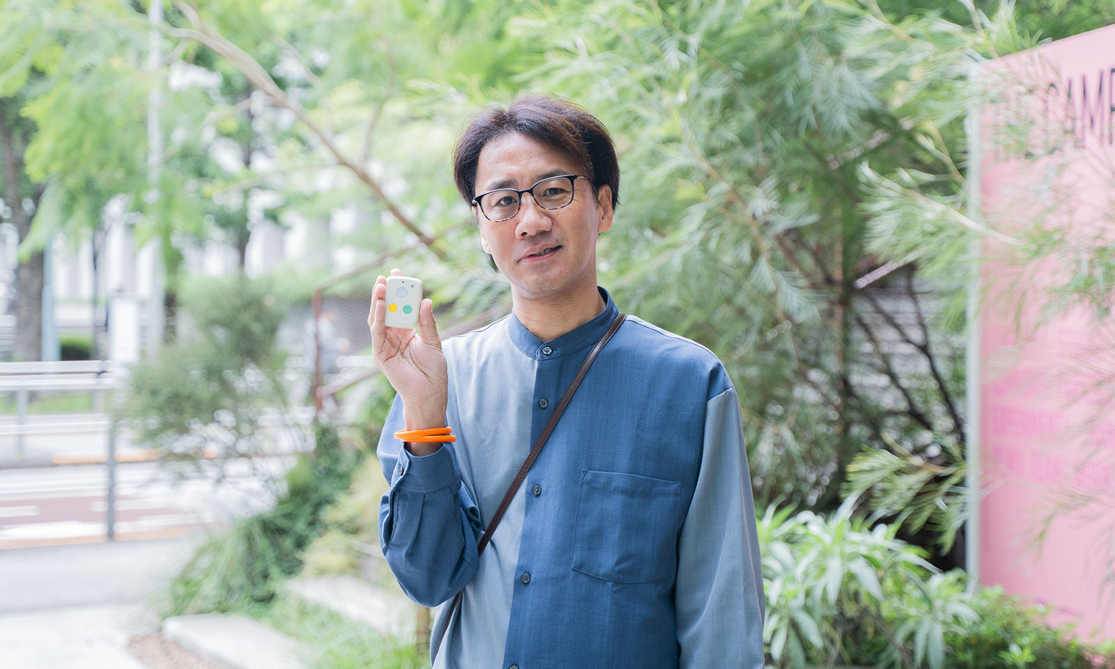 ALTはじまり。木々の前に立つ丹野智文さん。ブルーのシャツに、黒いメガネをかけ、手首には蛍光オレンジのリングを着用している。微笑みながら、ベージュ色の端末を持っている様子。ALTおわり。