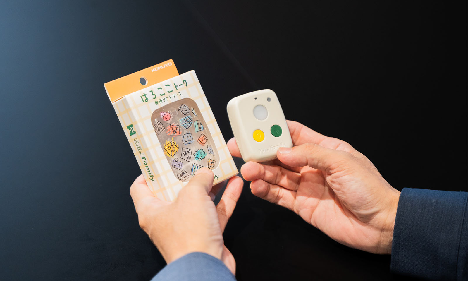 ALTはじまり。はろここトークの端末とパッケージを手に持っている。端末には、ベージュ色の角の丸い四角形の半透明の光るボタンの下に、黄色と緑のボタンがついている。ALTおわり。