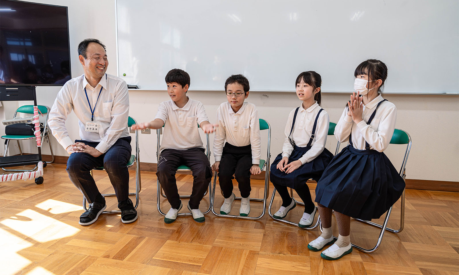 小学校の教室で、 改田先生と全校集会の企画担当となった4年生。一人の学生がおおきく手を広げ、ひょうきんな顔をしている。