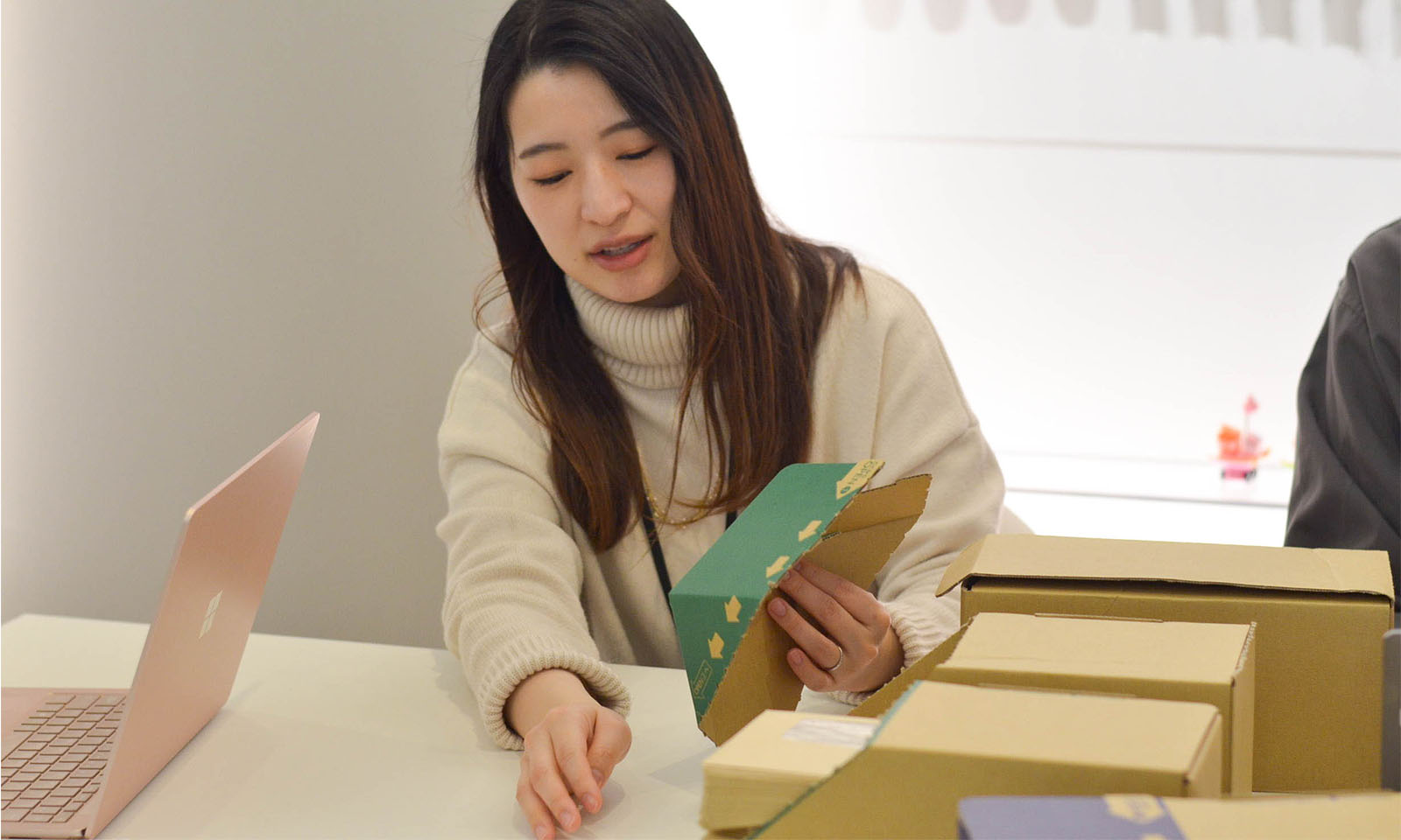 白いセーターを着たカウネット商品開発担当の小原(おはら)さんが左手に商品を手にとり、箱の形状を説明する様子