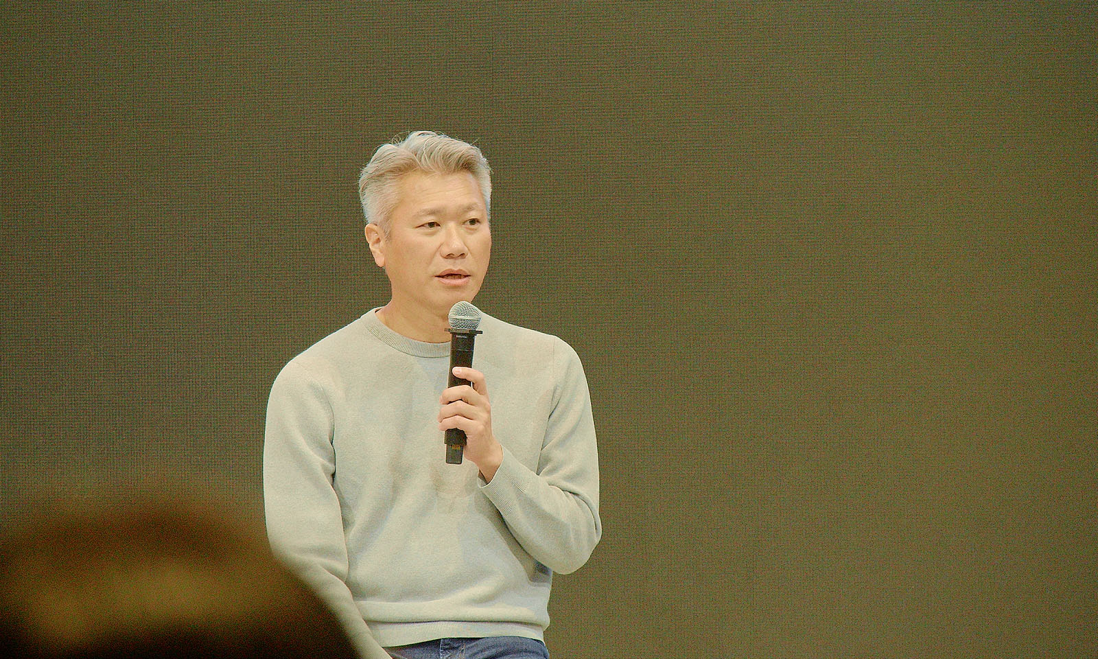 ALTはじまり。講演する越川さんの様子。薄いグレーのカットソーを着ている越川さんがマイクを持ち、聴衆に向かって話している。写真手間には聴衆の後頭部が見えている。ALTおわり