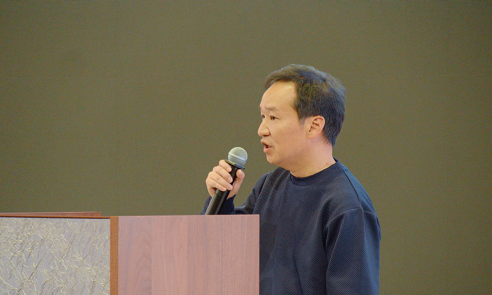 ALTはじまり。講演するコクヨKハート社長の西林さん。木製の司会者台の前に立ち、黒のカットソーを着た西林さんがマイクを持ち、聴衆に向かって話している。ALTおわり