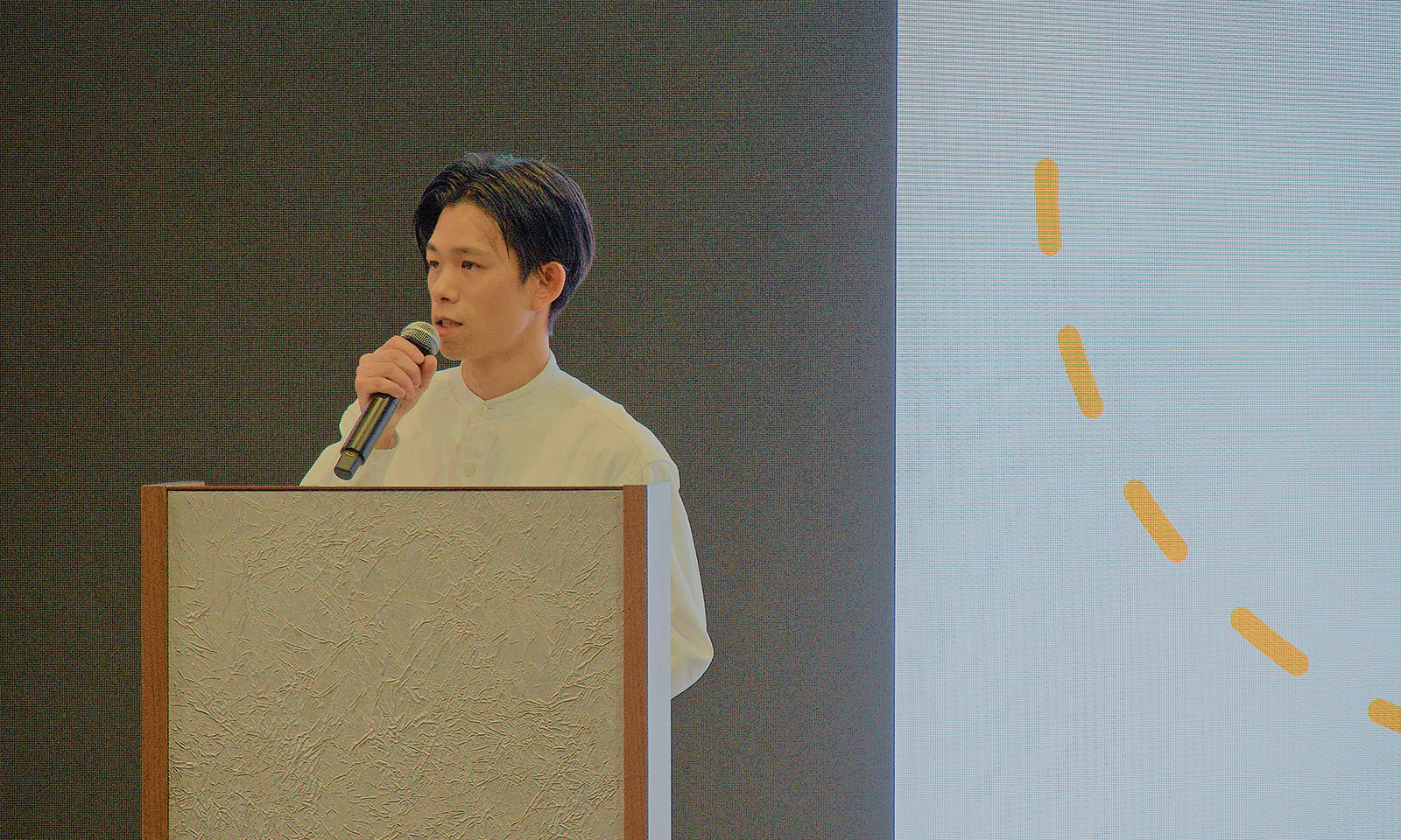 ALTはじまり。司会者台の前に立ち、マイクを持って講演する前田さん。清潔感のある白のシャツ姿で、背景にはプレゼン資料を映したスクリーンが写っている。ALTおわり