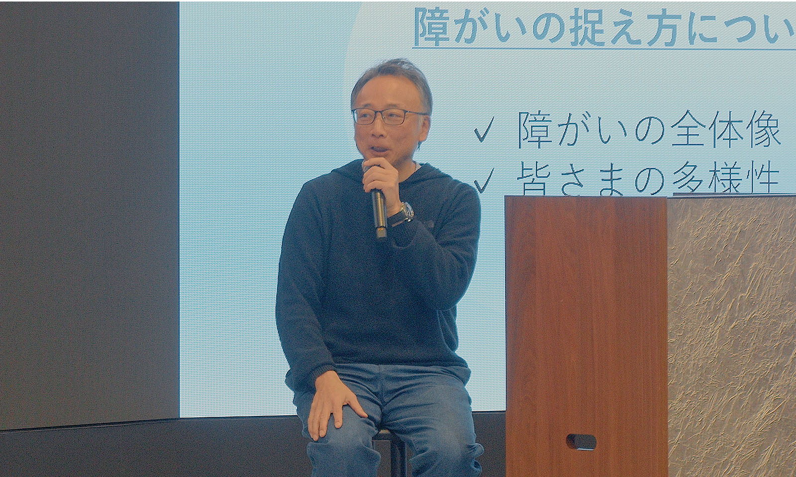 ALTはじまり。座談会の司会者を務めるコクヨの井田さん。黒のパーカーというカジュアルな服装で、左手にはマイクを持っている。ALTおわり